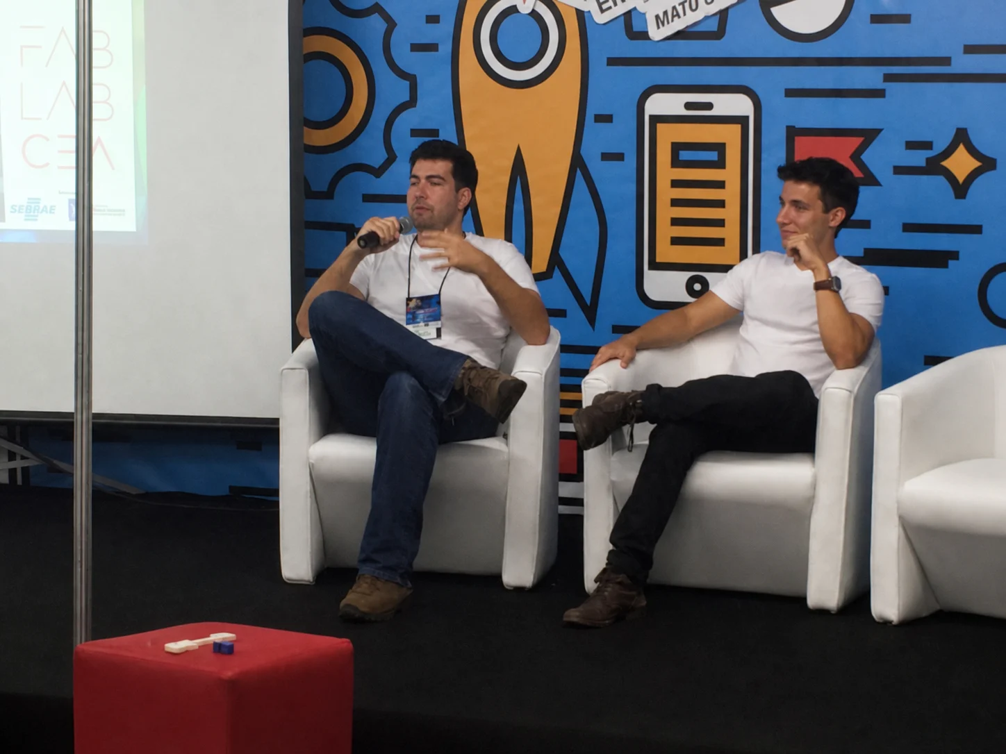 João participando de um painel com microfone ao lado de outro palestrante